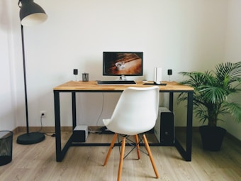 A minimalist workspace featuring a wooden desk with a black frame, holding a computer monitor and speakers. A white plastic chair with wooden legs is positioned in front of the desk, while a potted green plant and a black floor lamp flank the setup. The room has light wood flooring and white walls, lending a clean and modern aesthetic.