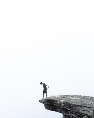 A person stands at the edge of a rock outcrop extending over a vast, misty landscape. The scene conveys a sense of solitude and introspection, with a minimalistic and serene backdrop.