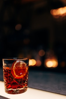 A crystal glass filled with a dark amber liquid and garnished with a slice of orange rests on a bar surface. The background is softly blurred with warm, ambient lighting creating a cozy, intimate atmosphere.