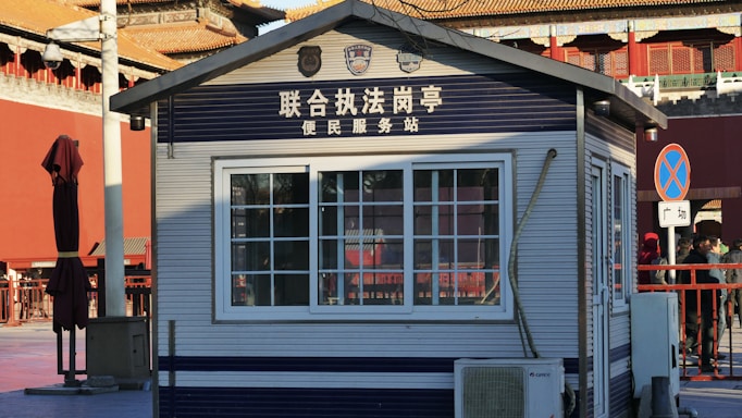 A small structure resembling a service or information kiosk, marked with Chinese text above the window and various logos. The building has a window with bars and air conditioning unit beneath it. Surrounding the kiosk is a red wooden umbrella stand and a few people on the side. The setting appears to be urban with visible traditional architectural elements in the background.