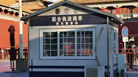 A small structure resembling a service or information kiosk, marked with Chinese text above the window and various logos. The building has a window with bars and air conditioning unit beneath it. Surrounding the kiosk is a red wooden umbrella stand and a few people on the side. The setting appears to be urban with visible traditional architectural elements in the background.