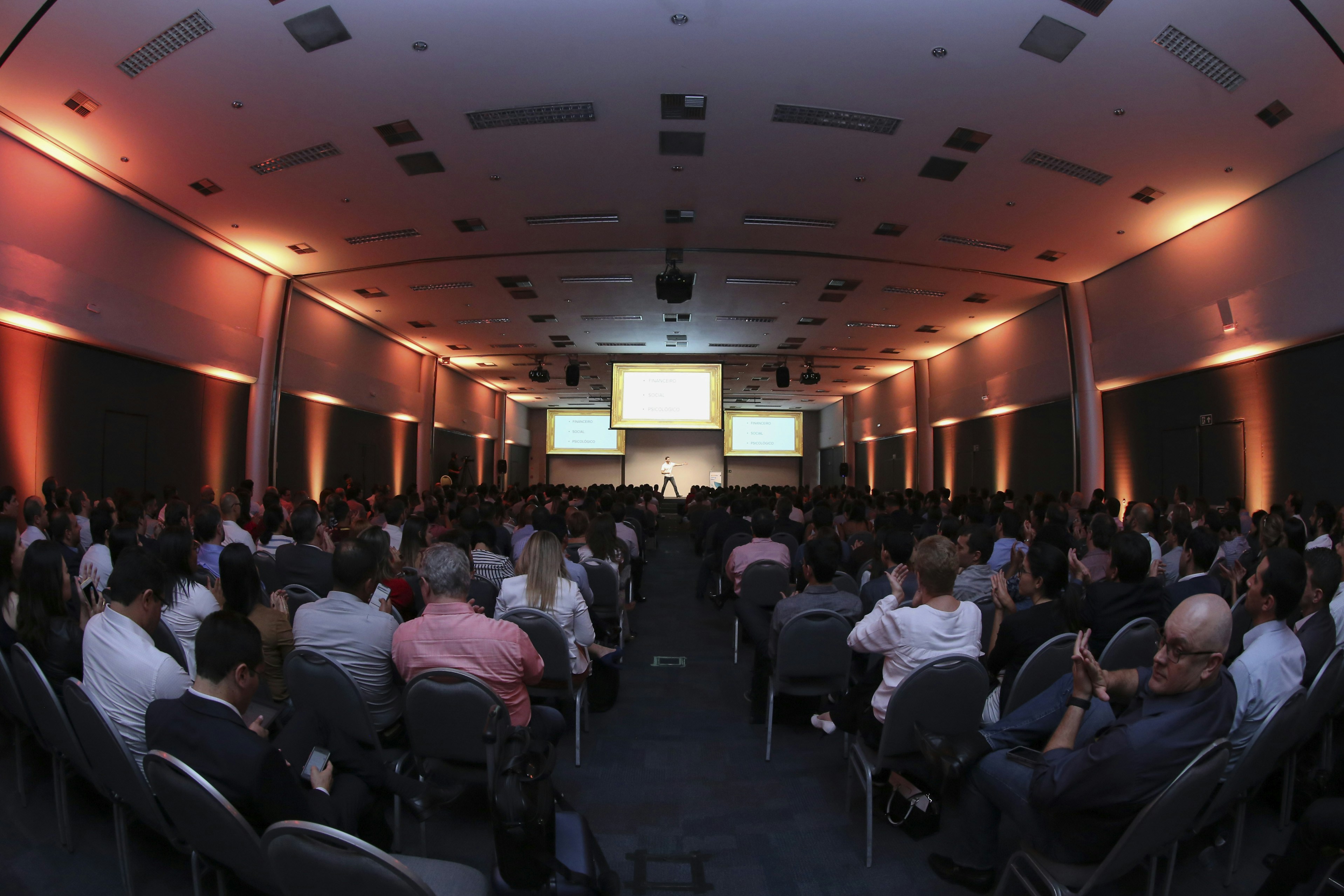A large conference room filled with people seated in rows, facing a stage with a presenter and several large screens displaying presentations. The lighting has a warm orange hue illuminating the walls, creating a professional and engaging atmosphere.