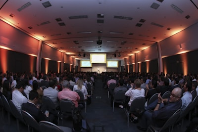 A large conference room filled with people seated in rows, facing a stage with a presenter and several large screens displaying presentations. The lighting has a warm orange hue illuminating the walls, creating a professional and engaging atmosphere.
