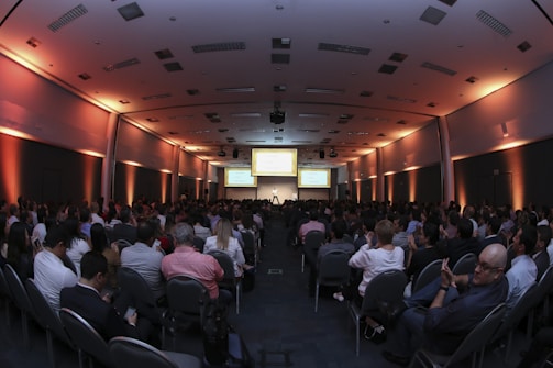 A large conference room filled with people seated in rows, facing a stage with a presenter and several large screens displaying presentations. The lighting has a warm orange hue illuminating the walls, creating a professional and engaging atmosphere.