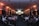 A large conference room filled with people seated in rows, facing a stage with a presenter and several large screens displaying presentations. The lighting has a warm orange hue illuminating the walls, creating a professional and engaging atmosphere.