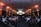 A large conference room filled with people seated in rows, facing a stage with a presenter and several large screens displaying presentations. The lighting has a warm orange hue illuminating the walls, creating a professional and engaging atmosphere.