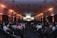 A large conference room filled with people seated in rows, facing a stage with a presenter and several large screens displaying presentations. The lighting has a warm orange hue illuminating the walls, creating a professional and engaging atmosphere.