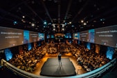 A large audience is seated in an auditorium with a speaker on stage. The room is dimly lit, with screens on either side displaying text and graphics related to the topic of management and work. The atmosphere suggests a professional conference or seminar.