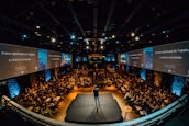 A large audience is seated in an auditorium with a speaker on stage. The room is dimly lit, with screens on either side displaying text and graphics related to the topic of management and work. The atmosphere suggests a professional conference or seminar.