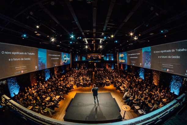 A large audience is seated in an auditorium with a speaker on stage. The room is dimly lit, with screens on either side displaying text and graphics related to the topic of management and work. The atmosphere suggests a professional conference or seminar.