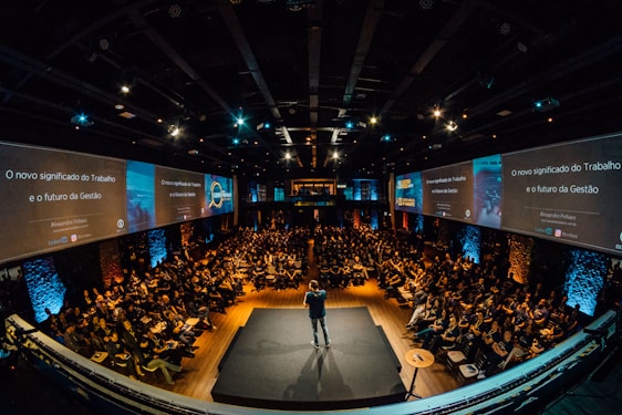 A large audience is seated in an auditorium with a speaker on stage. The room is dimly lit, with screens on either side displaying text and graphics related to the topic of management and work. The atmosphere suggests a professional conference or seminar.