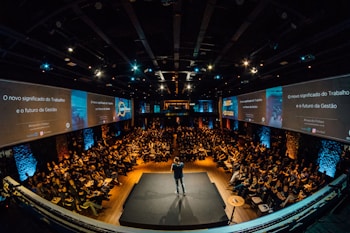 A large audience is seated in an auditorium with a speaker on stage. The room is dimly lit, with screens on either side displaying text and graphics related to the topic of management and work. The atmosphere suggests a professional conference or seminar.