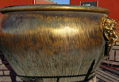 A large, ornate brass urn with a textured surface, featuring an intricate lion head design on the side holding a ring in its mouth. The urn is placed against a red backdrop, suggesting an outdoor setting, possibly in a historical or cultural site.