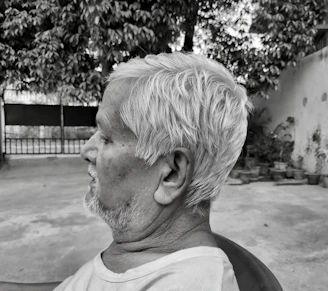 A side profile of an elderly man with short white hair. He is sitting outdoors on a chair, with a serene expression, in front of a backdrop of potted plants and a large tree. The scene has a grayscale tone, emphasizing textures and contrasts.
