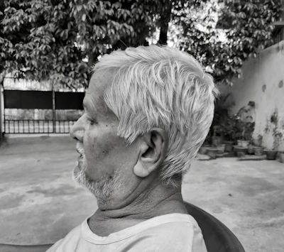 A side profile of an elderly man with short white hair. He is sitting outdoors on a chair, with a serene expression, in front of a backdrop of potted plants and a large tree. The scene has a grayscale tone, emphasizing textures and contrasts.