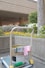 A cleaning cart is positioned in an outdoor area with a concrete building structure in the background. The cart holds a large bucket and cleaning supplies, including a striped cloth and a pink cloth hanging over the handle. A green scrubbing brush is placed on the bottom platform. Surrounding the area are vibrant flower beds with yellow and white flowers, as well as bushes and trees, contributing to a tidy and well-maintained environment.