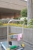 A cleaning cart is positioned in an outdoor area with a concrete building structure in the background. The cart holds a large bucket and cleaning supplies, including a striped cloth and a pink cloth hanging over the handle. A green scrubbing brush is placed on the bottom platform. Surrounding the area are vibrant flower beds with yellow and white flowers, as well as bushes and trees, contributing to a tidy and well-maintained environment.