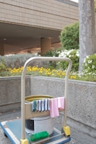 A cleaning cart is positioned in an outdoor area with a concrete building structure in the background. The cart holds a large bucket and cleaning supplies, including a striped cloth and a pink cloth hanging over the handle. A green scrubbing brush is placed on the bottom platform. Surrounding the area are vibrant flower beds with yellow and white flowers, as well as bushes and trees, contributing to a tidy and well-maintained environment.