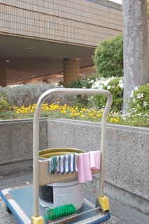 A cleaning cart is positioned in an outdoor area with a concrete building structure in the background. The cart holds a large bucket and cleaning supplies, including a striped cloth and a pink cloth hanging over the handle. A green scrubbing brush is placed on the bottom platform. Surrounding the area are vibrant flower beds with yellow and white flowers, as well as bushes and trees, contributing to a tidy and well-maintained environment.
