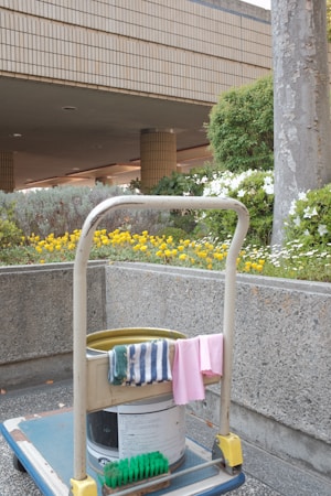 A cleaning cart is positioned in an outdoor area with a concrete building structure in the background. The cart holds a large bucket and cleaning supplies, including a striped cloth and a pink cloth hanging over the handle. A green scrubbing brush is placed on the bottom platform. Surrounding the area are vibrant flower beds with yellow and white flowers, as well as bushes and trees, contributing to a tidy and well-maintained environment.