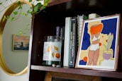 A wooden bookshelf holds a variety of items including a framed abstract artwork of a woman in a swimsuit, several books with visible titles like 'Cognitive Behavior Therapy', and a candle labeled 'Citrus & Cedarwood'. Some green leafy vines drape from above, and there's a golden-framed circular mirror reflecting part of the room.