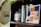 A wooden bookshelf holds a variety of items including a framed abstract artwork of a woman in a swimsuit, several books with visible titles like 'Cognitive Behavior Therapy', and a candle labeled 'Citrus & Cedarwood'. Some green leafy vines drape from above, and there's a golden-framed circular mirror reflecting part of the room.
