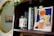 A wooden bookshelf holds a variety of items including a framed abstract artwork of a woman in a swimsuit, several books with visible titles like 'Cognitive Behavior Therapy', and a candle labeled 'Citrus & Cedarwood'. Some green leafy vines drape from above, and there's a golden-framed circular mirror reflecting part of the room.