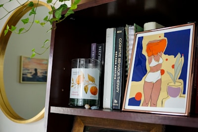 A wooden bookshelf holds a variety of items including a framed abstract artwork of a woman in a swimsuit, several books with visible titles like 'Cognitive Behavior Therapy', and a candle labeled 'Citrus & Cedarwood'. Some green leafy vines drape from above, and there's a golden-framed circular mirror reflecting part of the room.