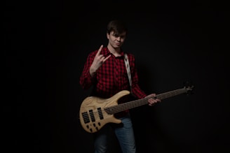 A person is standing against a dark background, wearing a red and black plaid shirt and jeans. They are holding an electric bass guitar, and using one hand to make a rock gesture. The instrument has a wooden finish with black pickups.