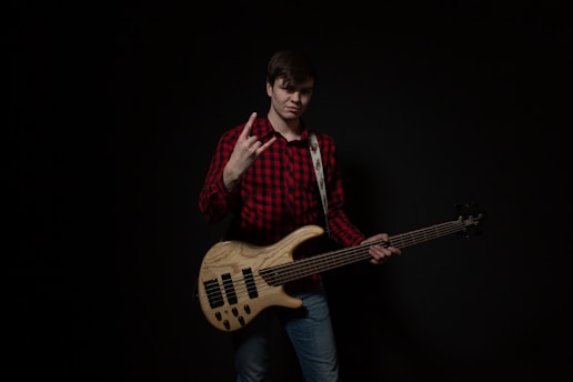 A person is standing against a dark background, wearing a red and black plaid shirt and jeans. They are holding an electric bass guitar, and using one hand to make a rock gesture. The instrument has a wooden finish with black pickups.