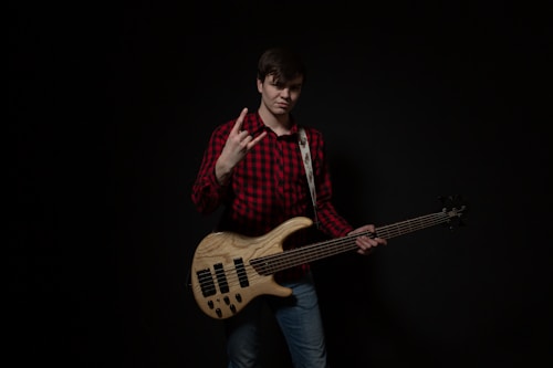 A person is standing against a dark background, wearing a red and black plaid shirt and jeans. They are holding an electric bass guitar, and using one hand to make a rock gesture. The instrument has a wooden finish with black pickups.