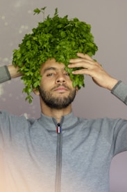 A person is humorously wearing a large bunch of green herbs as a wig on their head. They have a focused expression while holding the herbs with both hands. The person is wearing a gray zip-up sweater.