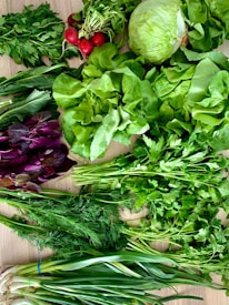 A variety of fresh vegetables and herbs are spread out on a wooden surface. The assortment includes leafy greens like lettuce, parsley, and dill, radishes with their tops still attached, green onions, and a head of cabbage. The vibrant colors and freshness of the produce suggest that it is ready for use in preparing meals.
