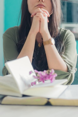 A woman with long dark hair is sitting with her hands clasped in front of her face, appearing to be in deep thought or prayer. She is wearing a green top and a watch on her left wrist. There is an open book with some visible text and a small bunch of purple flowers placed on the pages in front of her. The background is out of focus with a mix of green and other muted colors.