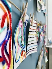 Colorful strings of yarn are attached to pieces of paper using clothespins, all hung on a rope against a blue wall. The strings are arranged in various patterns, providing a textured and vibrant display.
