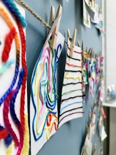 Colorful strings of yarn are attached to pieces of paper using clothespins, all hung on a rope against a blue wall. The strings are arranged in various patterns, providing a textured and vibrant display.