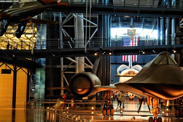 A museum exhibit featuring several historic aircrafts, including a space shuttle and a sleek, black jet. Surrounding the aircrafts are walkways and railings, with various visitors observing the displays. An American flag hangs prominently in the background, suggesting a space or aviation theme.