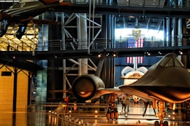 A museum exhibit featuring several historic aircrafts, including a space shuttle and a sleek, black jet. Surrounding the aircrafts are walkways and railings, with various visitors observing the displays. An American flag hangs prominently in the background, suggesting a space or aviation theme.