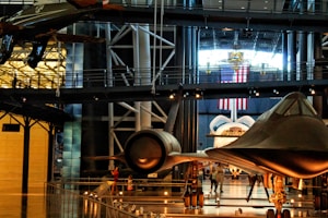 A museum exhibit featuring several historic aircrafts, including a space shuttle and a sleek, black jet. Surrounding the aircrafts are walkways and railings, with various visitors observing the displays. An American flag hangs prominently in the background, suggesting a space or aviation theme.