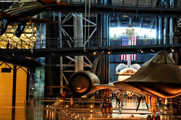 A museum exhibit featuring several historic aircrafts, including a space shuttle and a sleek, black jet. Surrounding the aircrafts are walkways and railings, with various visitors observing the displays. An American flag hangs prominently in the background, suggesting a space or aviation theme.