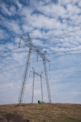Several large electricity pylons stand on a grassy hill beneath a partly cloudy sky. A small green van is parked near the base of the closer pylon.