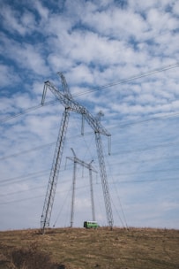 Several large electricity pylons stand on a grassy hill beneath a partly cloudy sky. A small green van is parked near the base of the closer pylon.