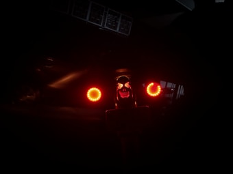 A dimly lit scene with two bright red circular lights resembling eyes, and a sinister face illuminated by red light. The background is mostly dark, with the faint outline of a vehicle and some barely visible structures.