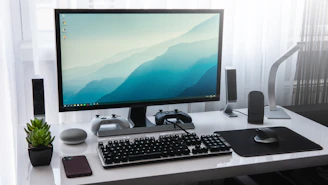 A computer monitor, keyboard, and game controllers on a white desk in a workspace