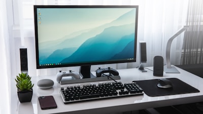 A modern computer setup on a white desk, featuring a widescreen monitor displaying a blue gradient wallpaper. Two gaming controllers are placed on stands underneath the monitor. A mechanical keyboard and a wireless mouse sit in front of the monitor on a mouse pad. A smartphone is lying next to a small potted plant on the left, and there is a smart speaker nearby. The desk is flanked by a sleek desk lamp and two speakers, with sheer white curtains in the background.