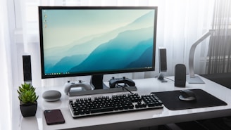 A modern computer setup on a white desk, featuring a widescreen monitor displaying a blue gradient wallpaper. Two gaming controllers are placed on stands underneath the monitor. A mechanical keyboard and a wireless mouse sit in front of the monitor on a mouse pad. A smartphone is lying next to a small potted plant on the left, and there is a smart speaker nearby. The desk is flanked by a sleek desk lamp and two speakers, with sheer white curtains in the background.