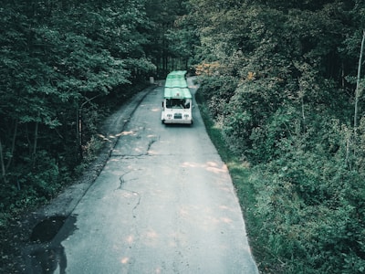 A white vehicle is driving along a narrow, winding road surrounded by dense green foliage. The vehicle has a green canoe strapped to its roof, indicating preparation for an outdoor adventure. The road is narrow and slightly cracked, flanked by lush trees that create a serene and secluded atmosphere.