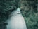 A white vehicle is driving along a narrow, winding road surrounded by dense green foliage. The vehicle has a green canoe strapped to its roof, indicating preparation for an outdoor adventure. The road is narrow and slightly cracked, flanked by lush trees that create a serene and secluded atmosphere.