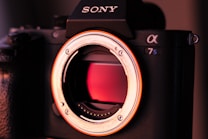 A close-up image of a Sony camera body without a lens attached. The camera features a prominent sensor visible through the lens mount. The orange and red lighting creates a warm atmosphere, highlighting the inner details of the camera.