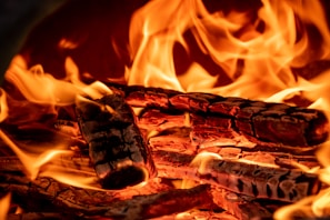 Burning logs with bright orange and yellow flames surrounding them. The wood appears charred and the flames are dynamic, with varying intensities and directions.